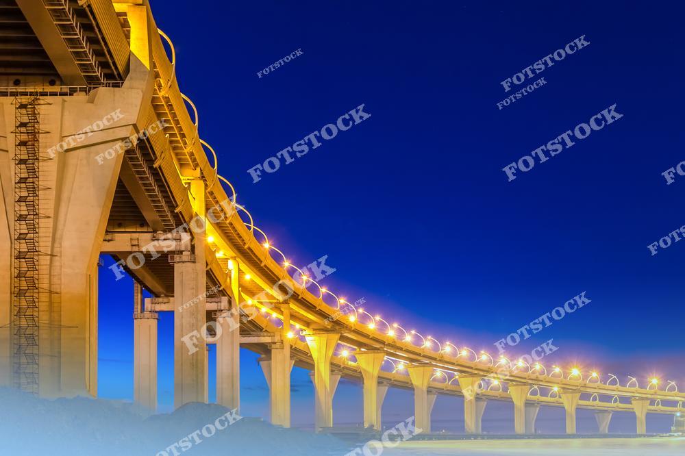 На фотографии запечатлен участок Западного скоростного диаметра (ЗСД) в Санкт-Петербурге в вечернее время. Мост, подсвеченный теплым светом фонарей, эффектно выделяется на фоне глубокого синего неба. Архитектурные линии ЗСД подчёркивают изогнутую форму трассы, которая соединяет разные части города. Этот современный инженерный объект стал важной частью городской инфраструктуры и символом технологического прогресса.